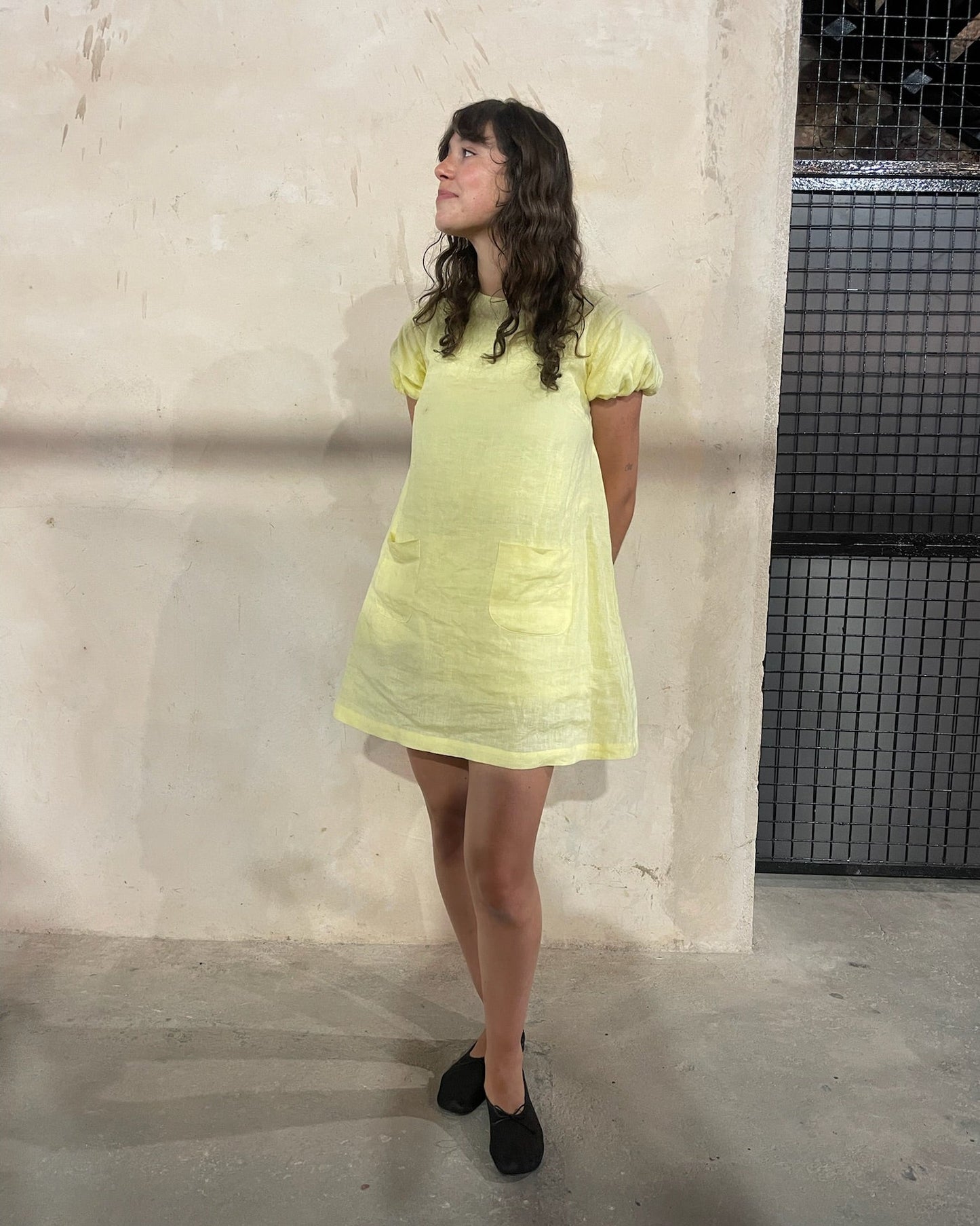 Woman wearing a light yellow dress standing against a plain wall.