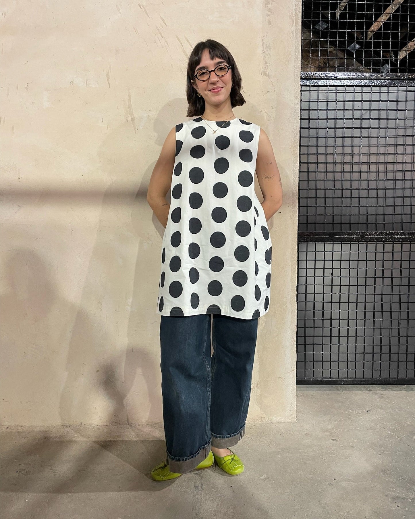 Person wearing a sleeveless polka dot dress and jeans standing against a plain wall.