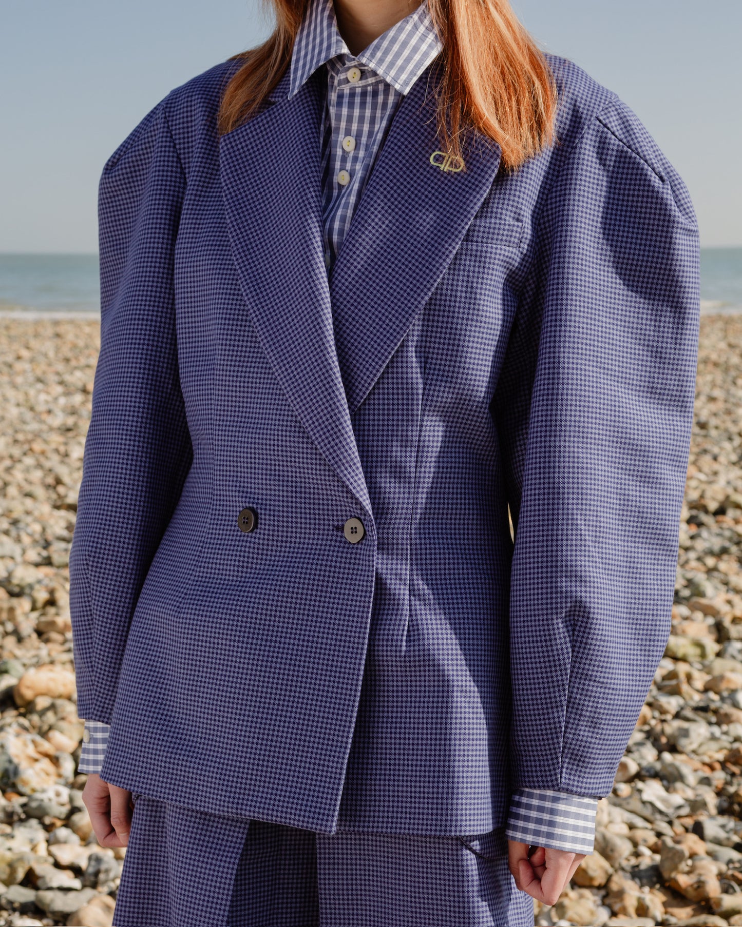 Person wearing a blue checkered blazer standing on a pebbly beach.