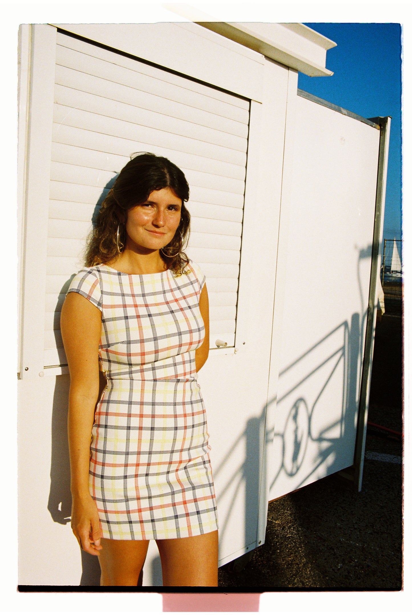 Woman in a plaid dress standing in front of a white building with a blue sky.