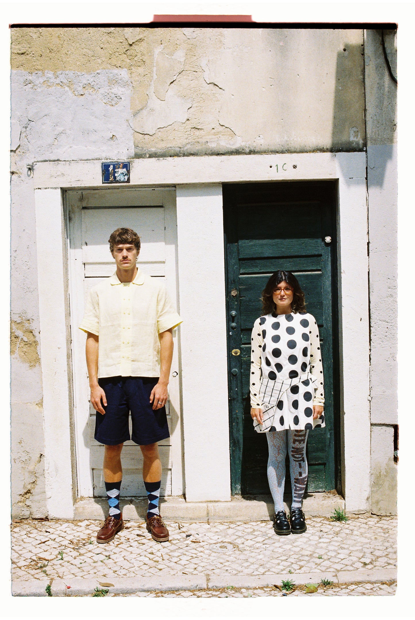 Two people standing in front of a building, one wearing polka dots and the other in yellow