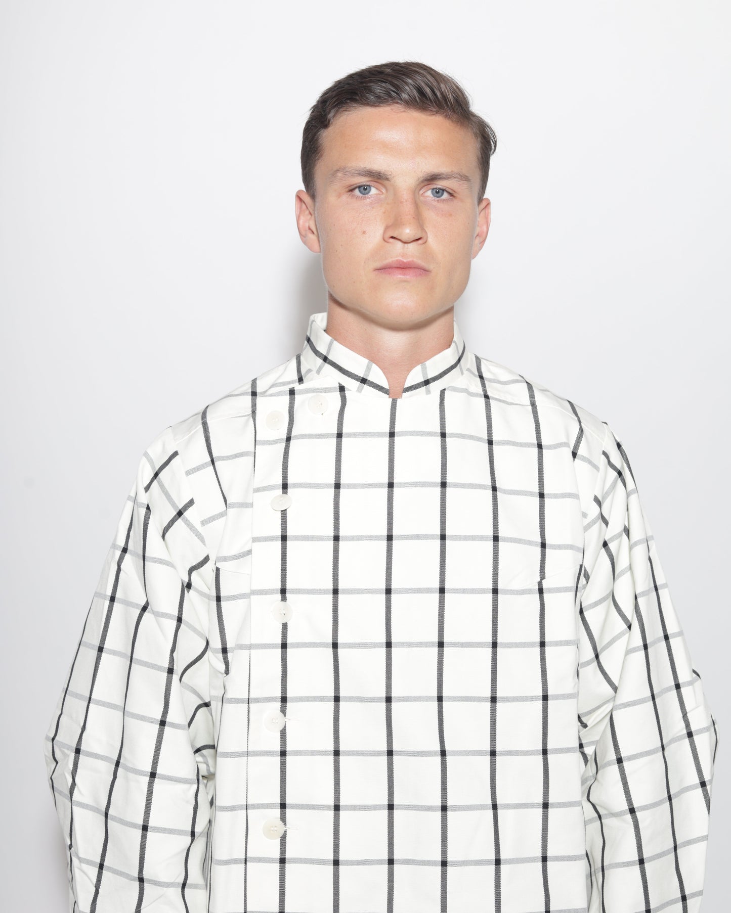 A portrait-style studio photograph of a man with short brown hair and blue eyes, wearing a white and black plaid jacket with a mandarin collar. The image is a close-up, showing his head and shoulders against a light background.