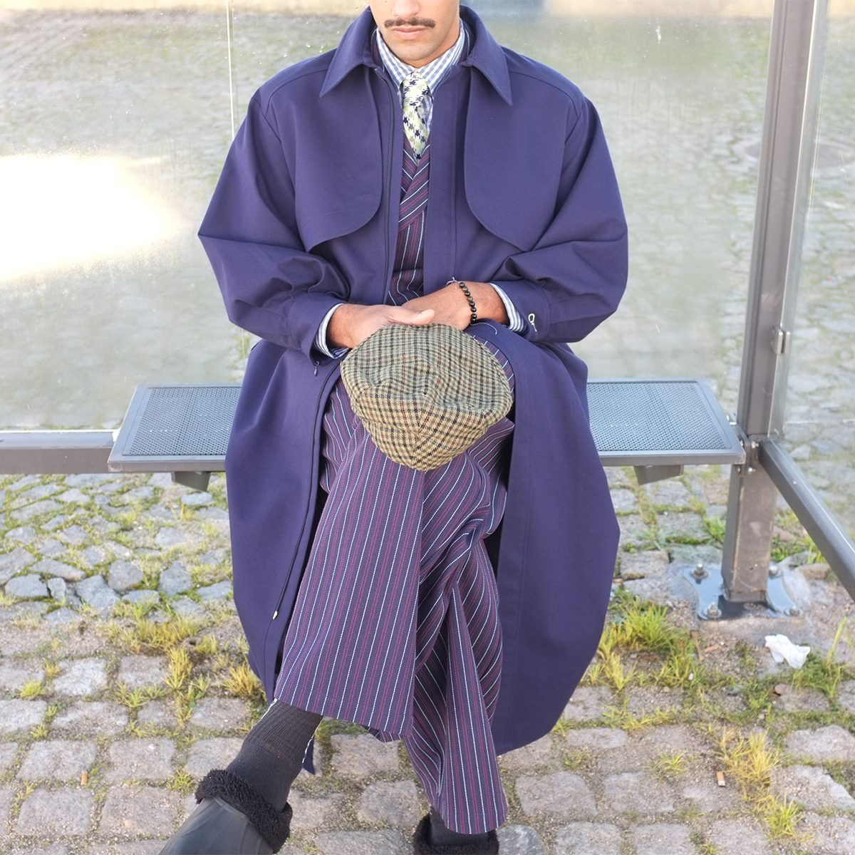 Person wearing a long blue coat and striped pants sitting on a bench.