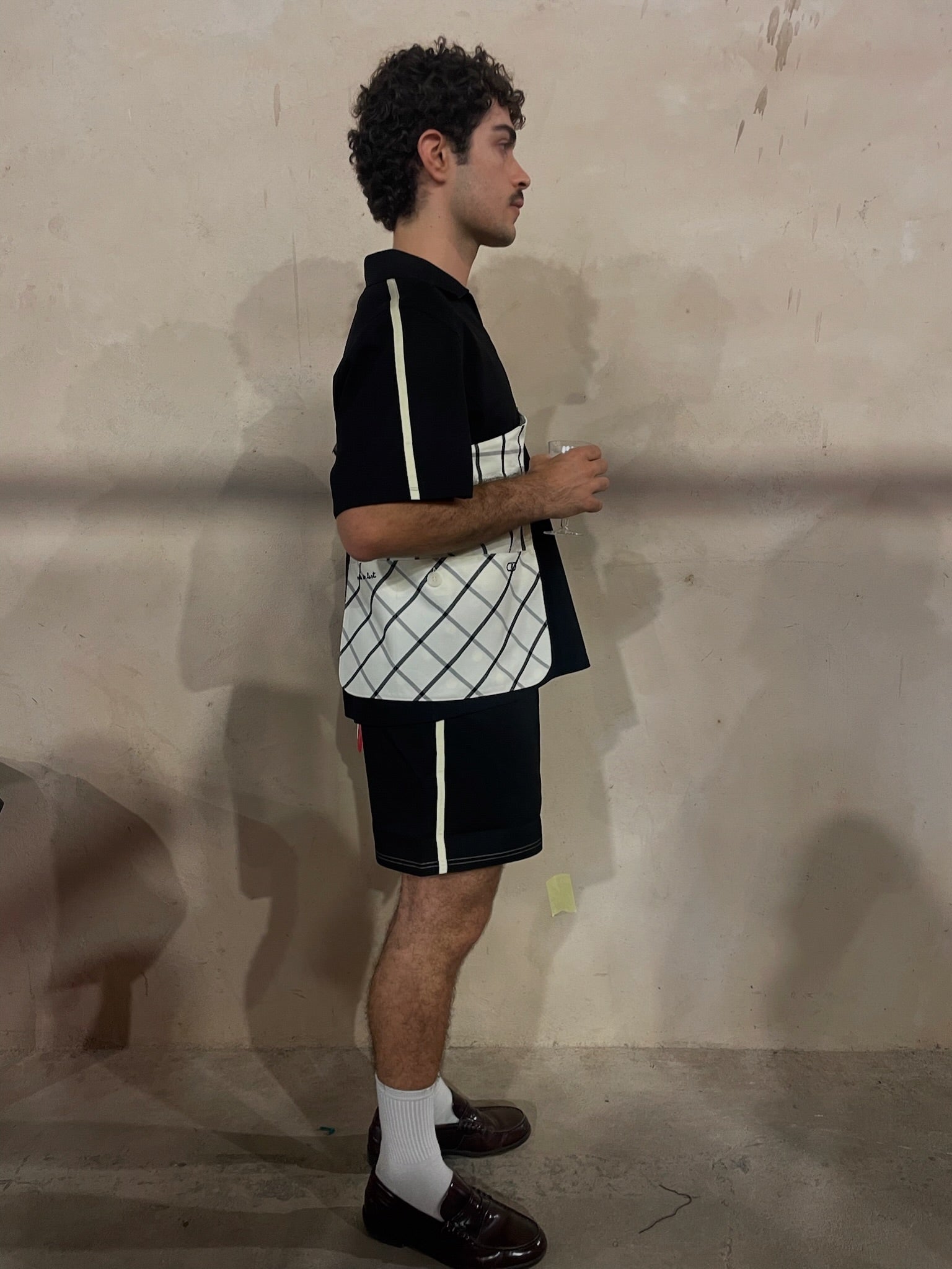 Man in a black polo and black matching shorts with a patterned bag against a plain wall