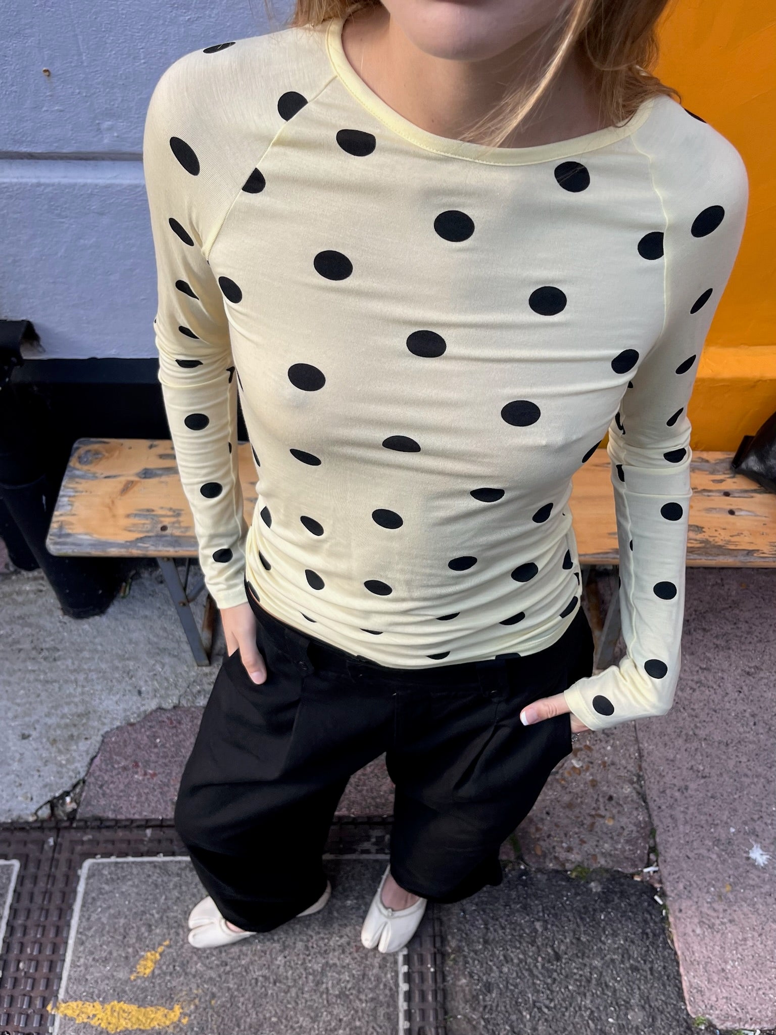 Person wearing a yellow long-sleeve top with black polka dots and black pants on a sidewalk.