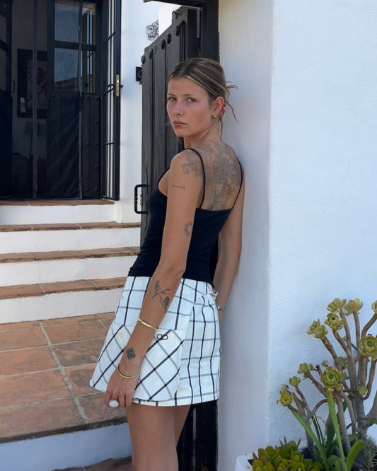 Woman leaning against a white wall with tattoos visible, wearing a black tank top and white checkered skirt.