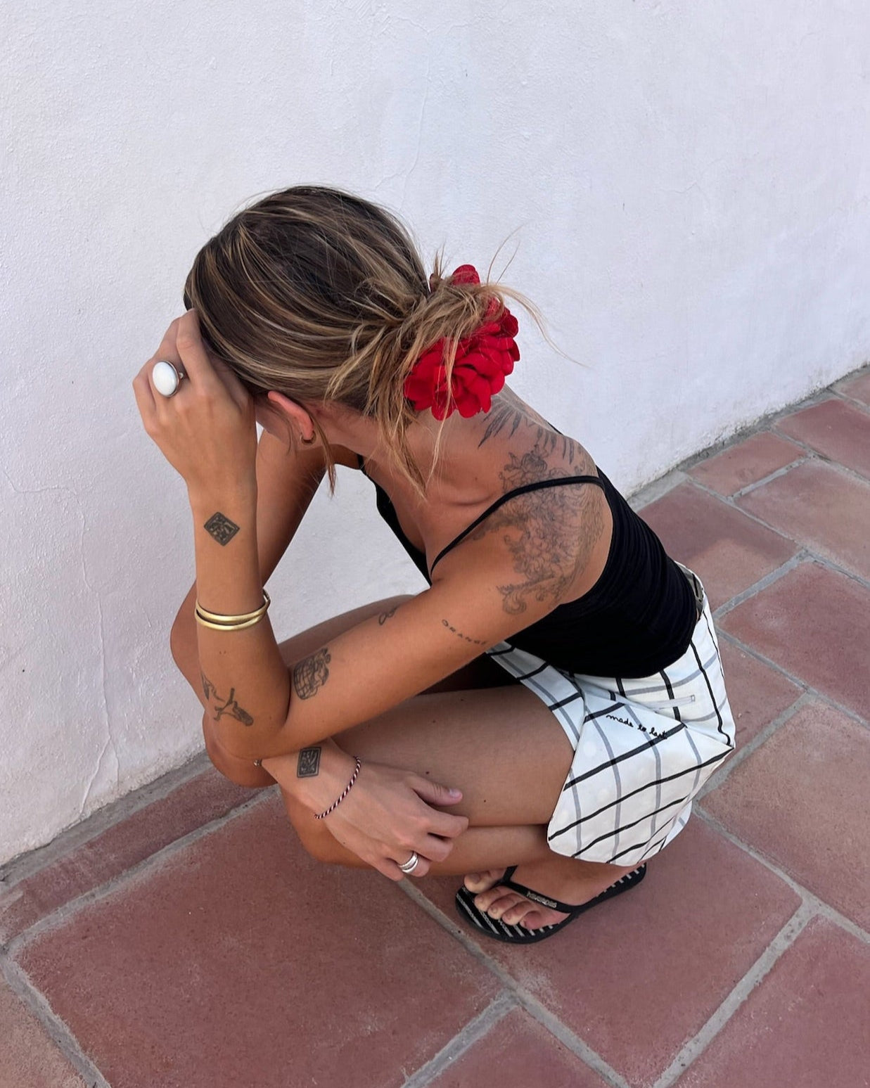Woman squatting on a tiled floor with a white wall background with a checkered skirt 