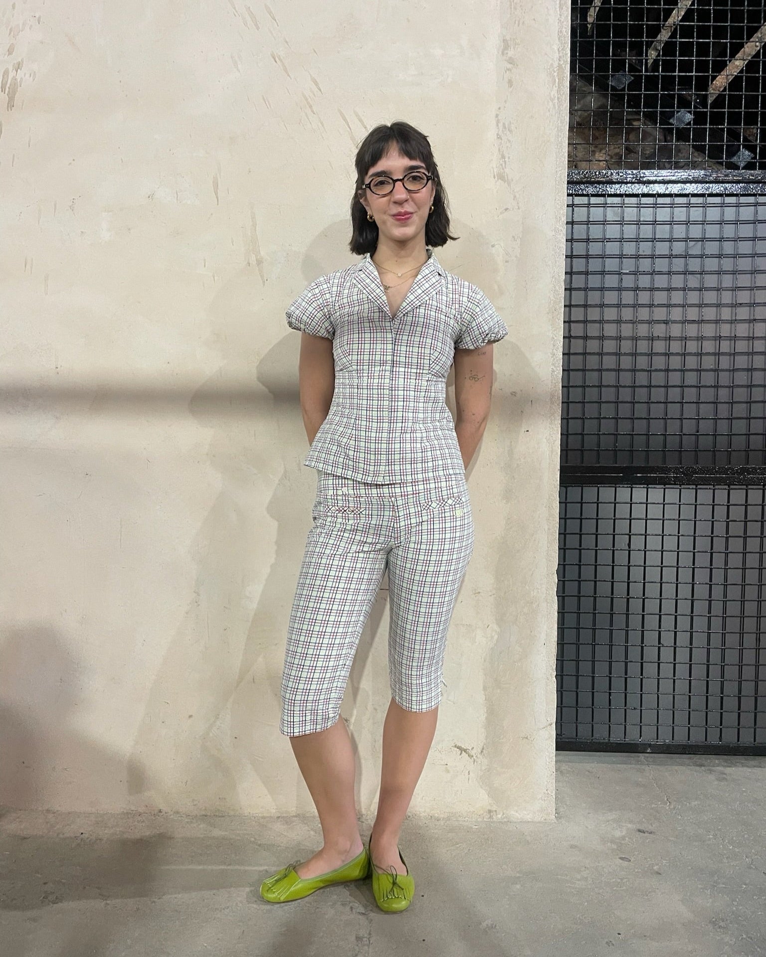 Person wearing a checkered blouse and matching capris standing against a textured wall.