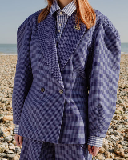 Person wearing a blue checkered blazer standing on a pebbly beach.