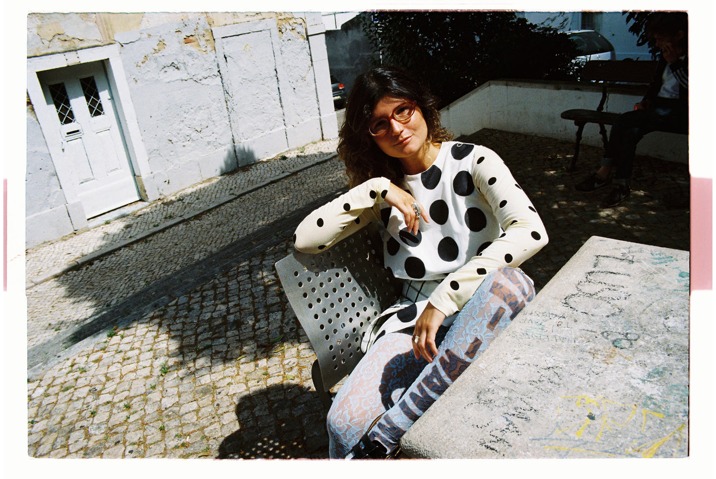Woman sitting on a bench wearing a polka dot outfit with a white building and door in the background.