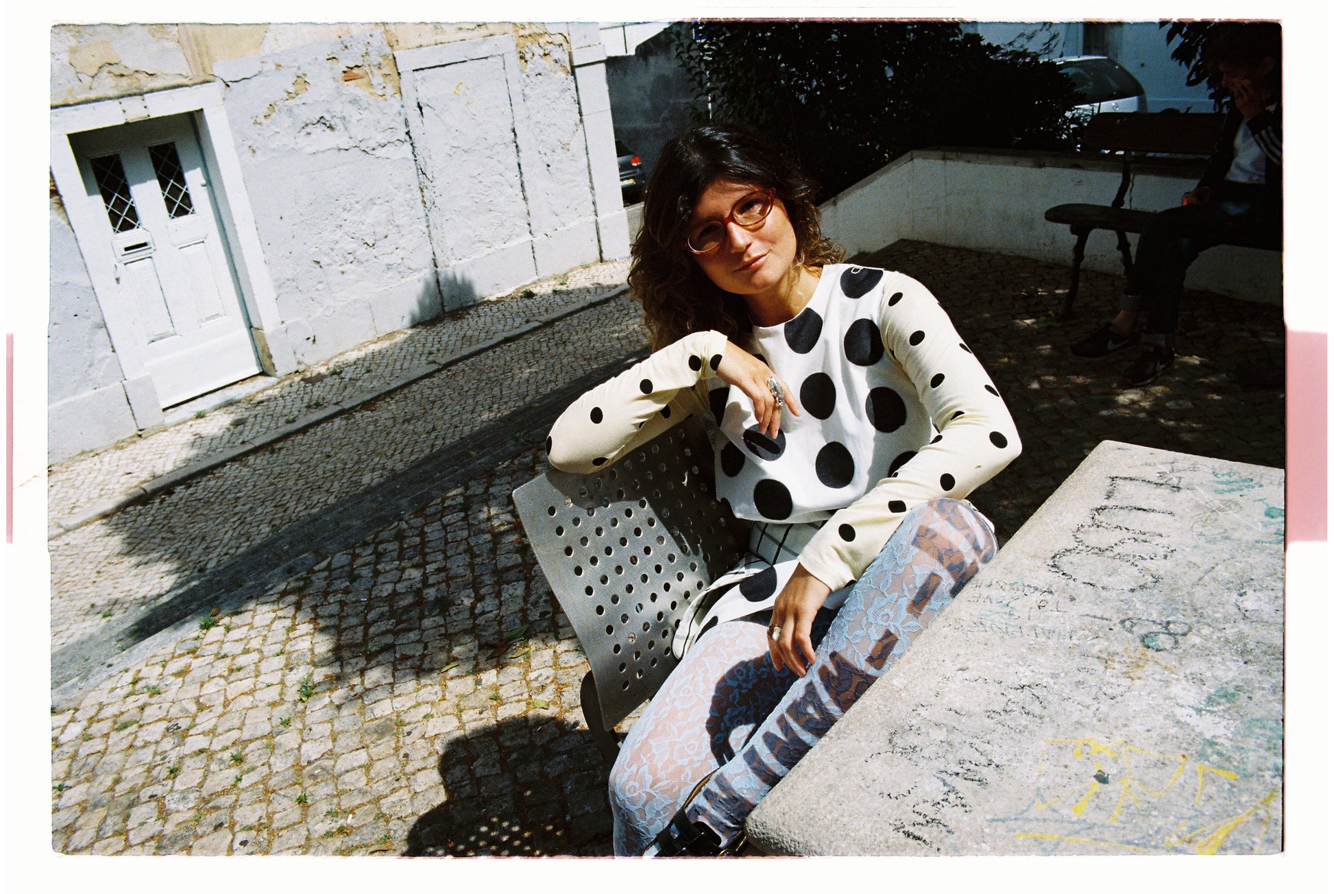Woman sitting on a bench wearing a polka dot outfit with a white building and door in the background.