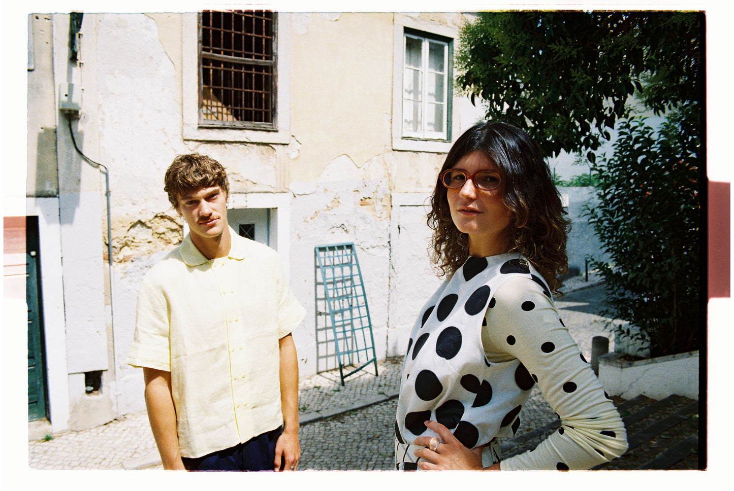 Two people standing outdoors with a building and trees in the background. One wearing polka dots and the other in a yellow shirt 