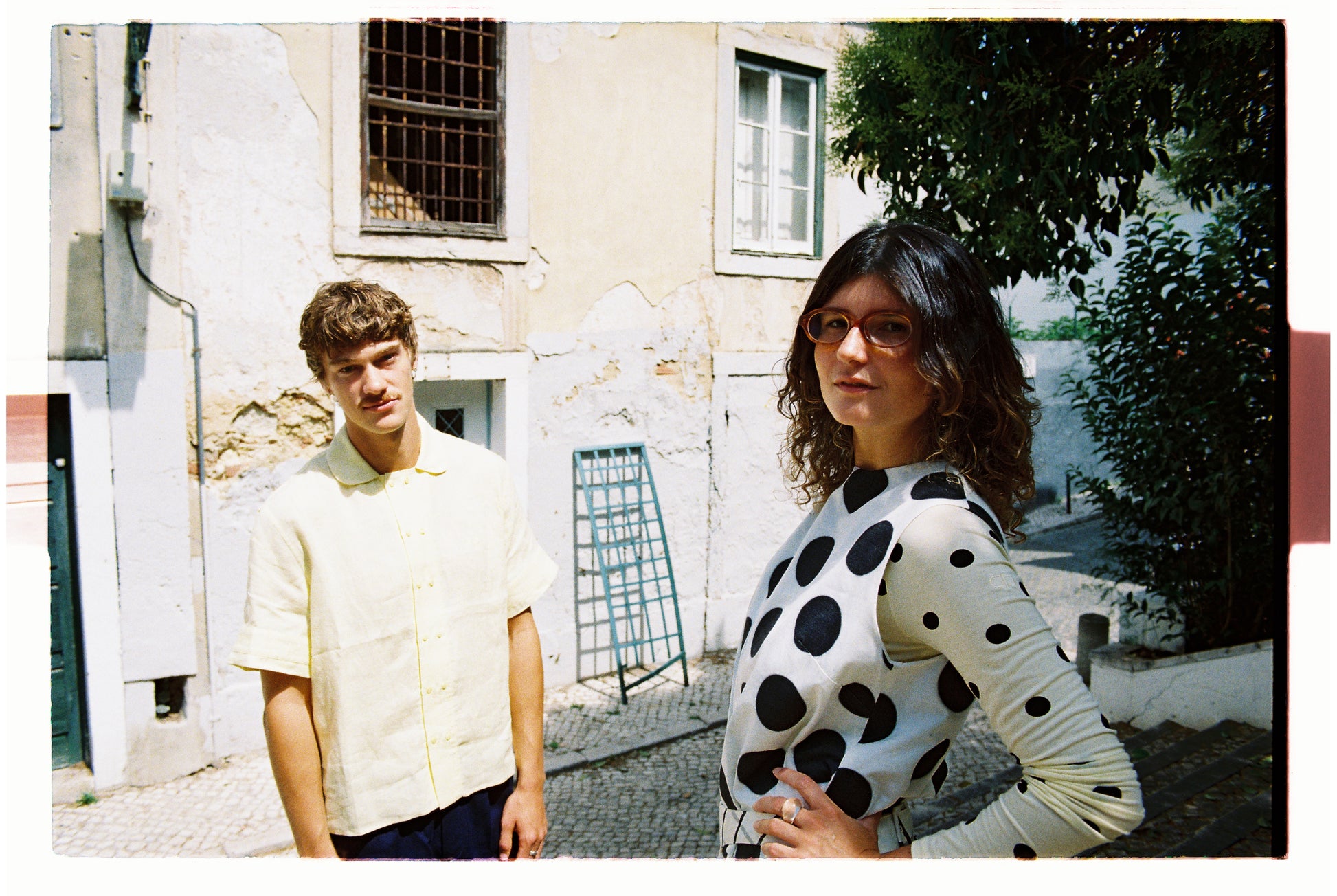 Two people standing outdoors with a building and trees in the background. One wearing polka dots and the other in a yellow shirt 