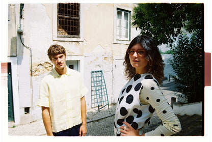 Two people standing outdoors with a building and trees in the background. One wearing polka dots and the other in a yellow shirt 