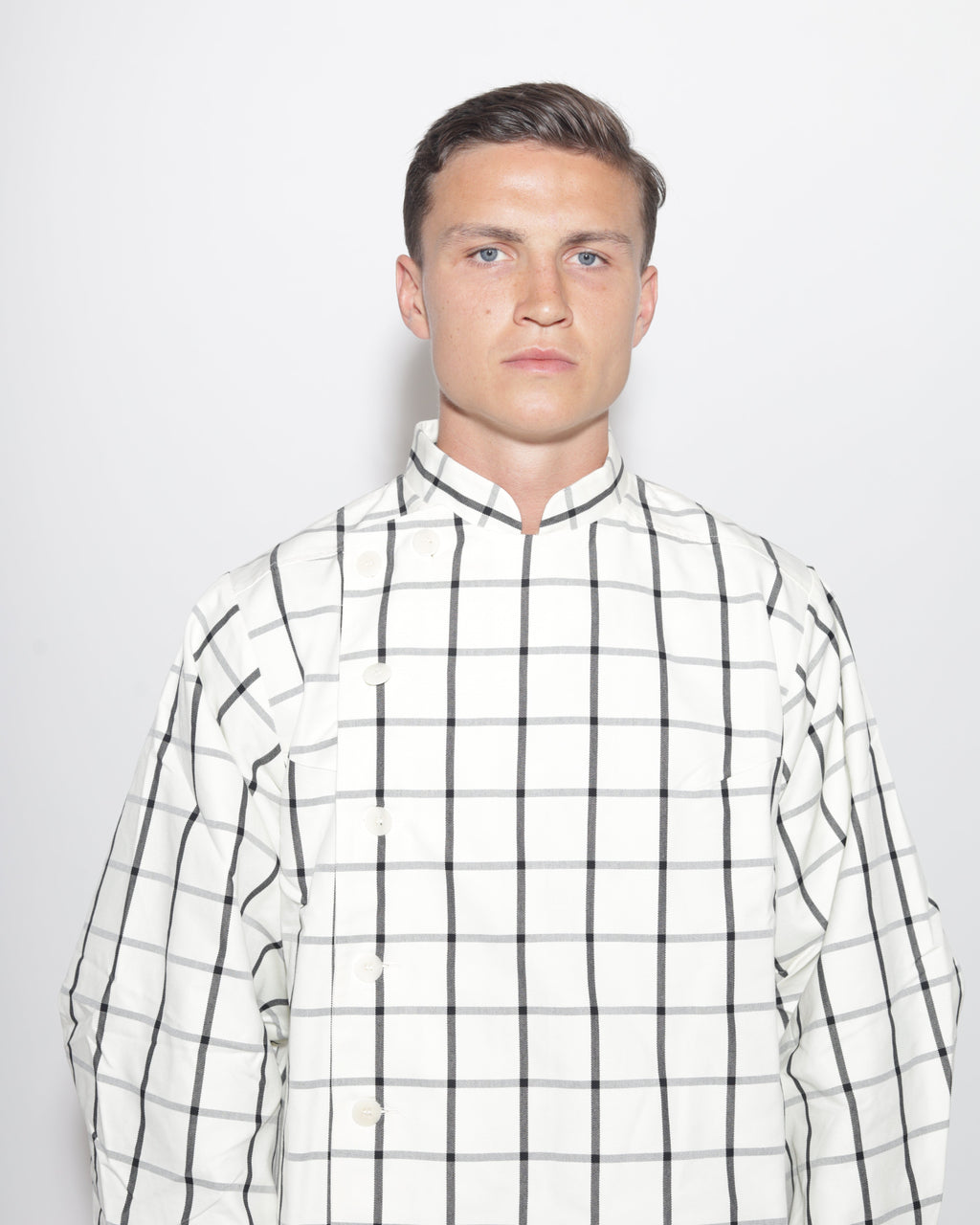 A portrait-style studio photograph of a man with short brown hair and blue eyes, wearing a white and black plaid jacket with a mandarin collar. The image is a close-up, showing his head and shoulders against a light background.