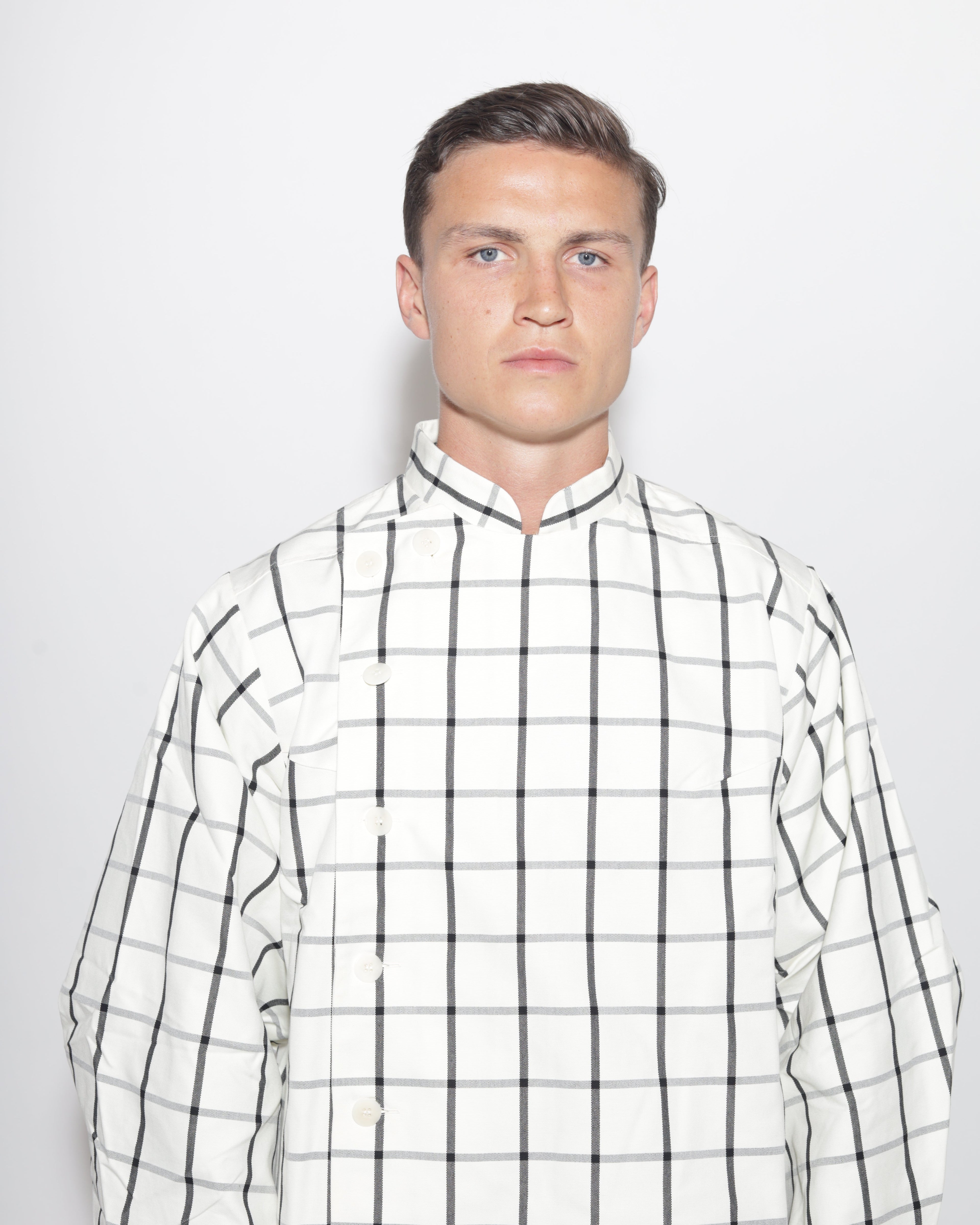 A portrait-style studio photograph of a man with short brown hair and blue eyes, wearing a white and black plaid jacket with a mandarin collar. The image is a close-up, showing his head and shoulders against a light background.